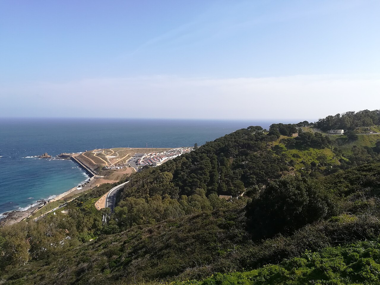 Vista panorámica de la costa de Ceuta desde una ladera arbolada, con el mar, un tramo de carretera y una zona edificada junto al espigón