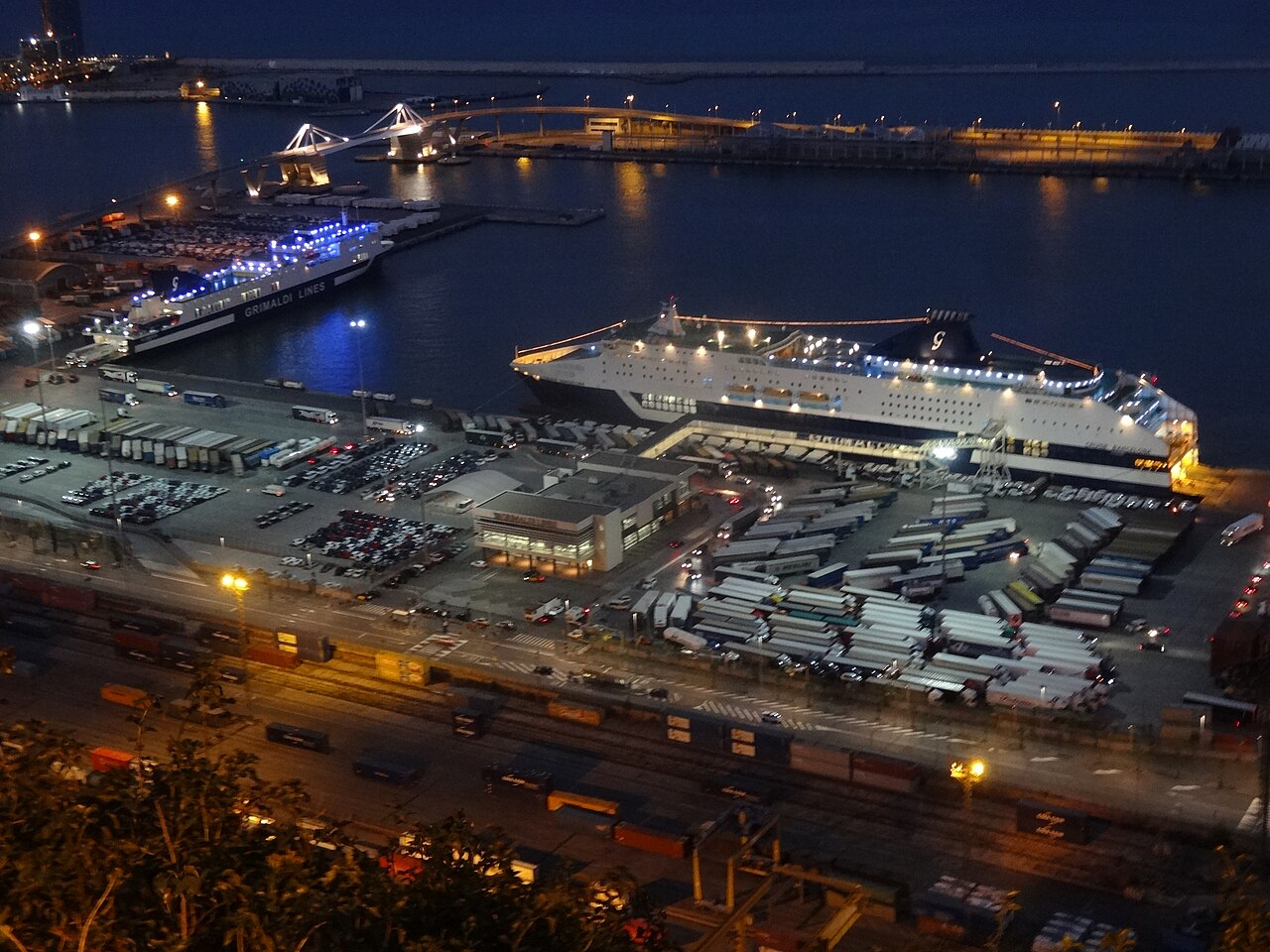 Vista nocturna de la terminal de ferris de pasajeros del puerto de Barcelona, con un buque de línea atracado y las playas de camiones y vehículos de la zona portuaria