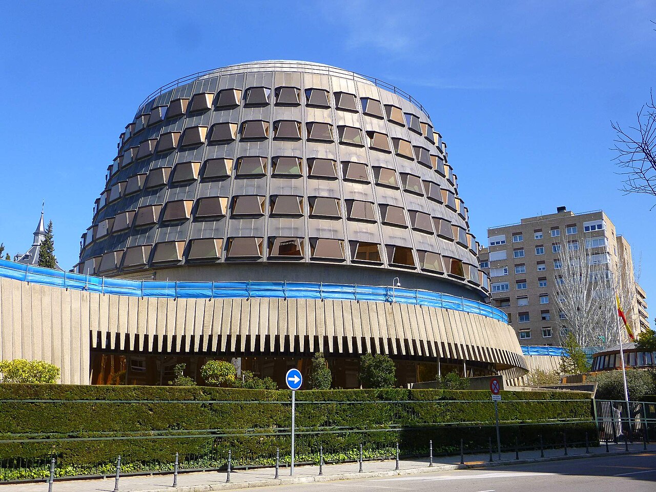 Fachada del edificio circular de la sede del Tribunal Constitucional en Madrid, con su cuerpo superior acristalado de ventanas cuadradas y el seto de la calle en primer término